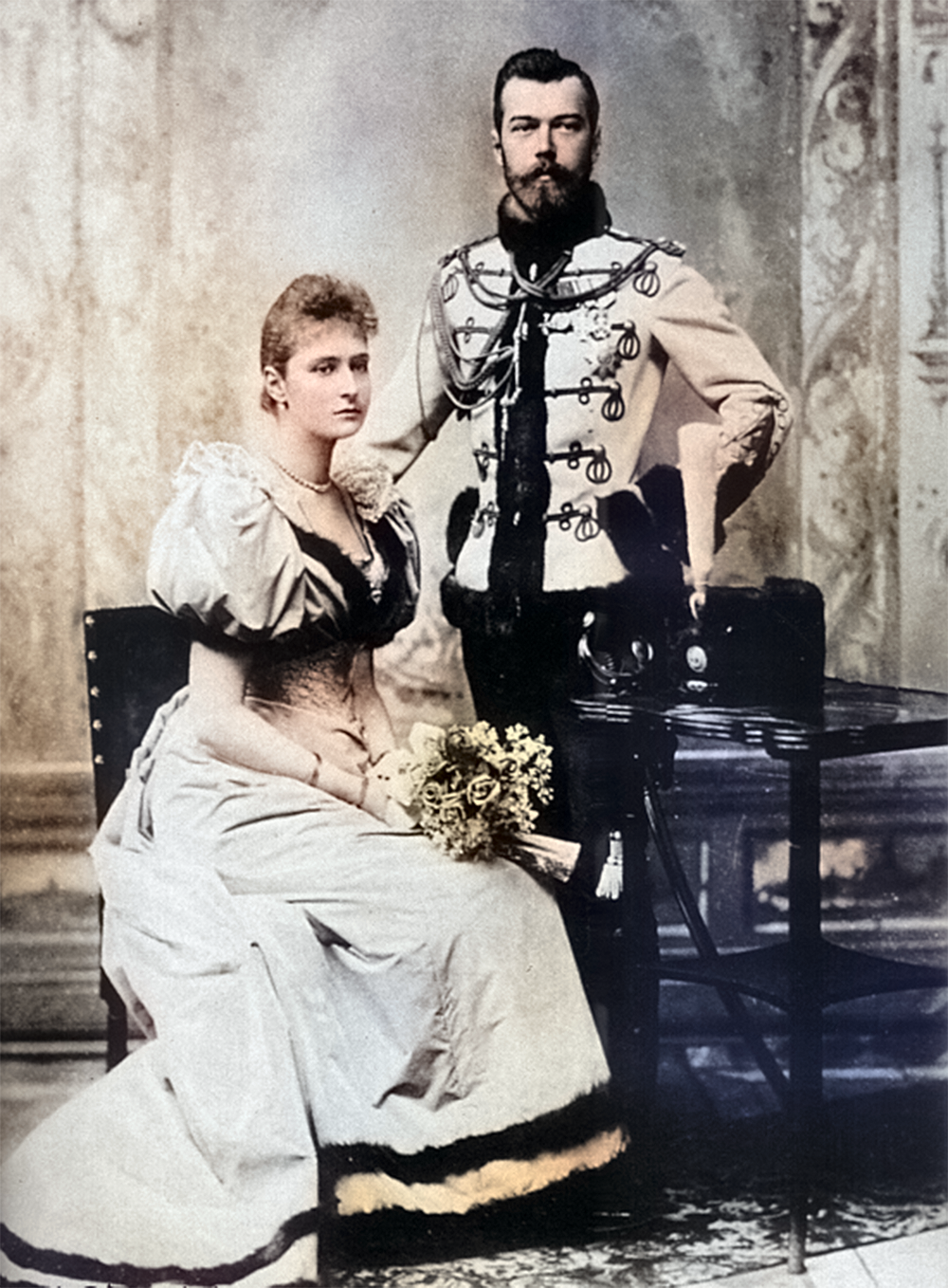 This is a colorized photograph of two people sitting in a formal setting. The man is wearing a elaborate formal suit and the woman is wearing a long dress with a train. The man has a stern expression on his face and the woman looks very worried.