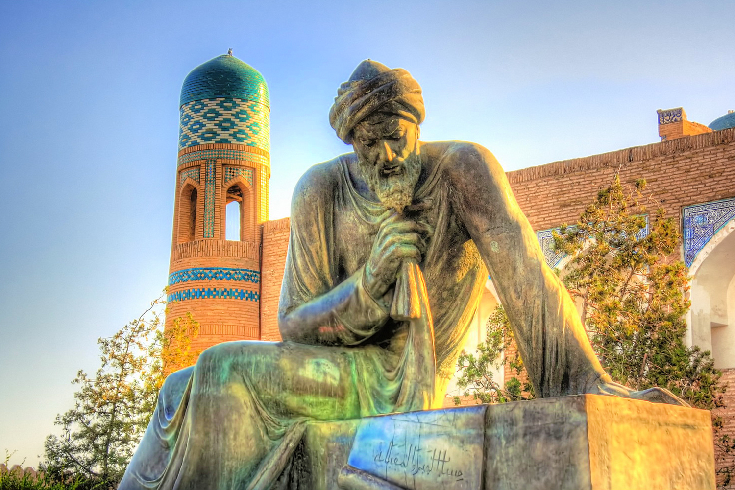 A photo of a bronze statue of Al-Khwarizmi, a Persian mathematician and astronomer, sitting with his head resting on his hand, in front of a blue-tiled tower at sunset.