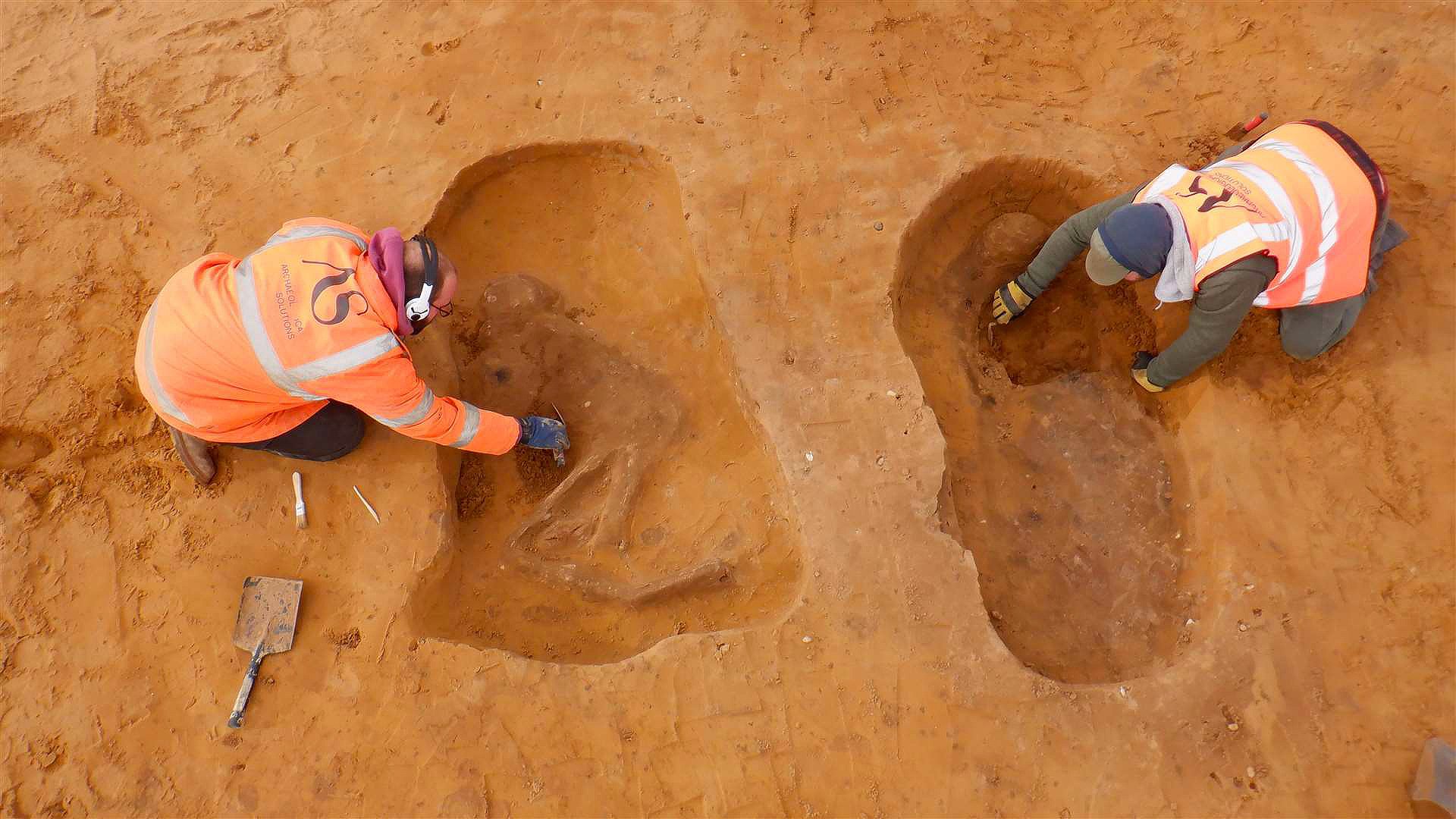 An aerial photo of two workers excavating two Anglo-Saxon graves in a red dirt area. The workers are wearing orange vests and hard hats, and there is a shovel and a pickaxe next to one of the graves.