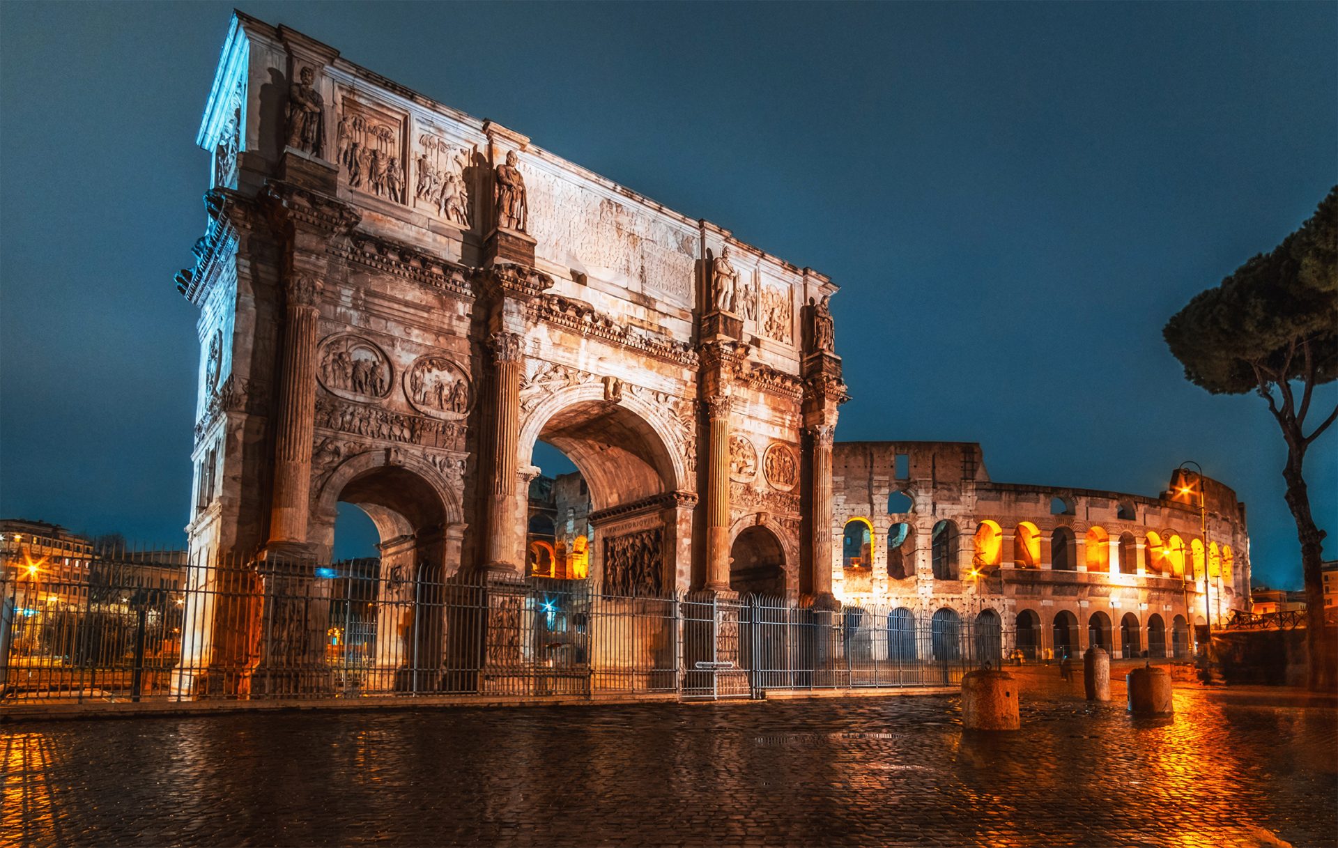 A photo of the Arch of Constantine and the Colosseum in Rome, Italy at night, with warm lights, a cloudy sky, and a cobblestone street.
