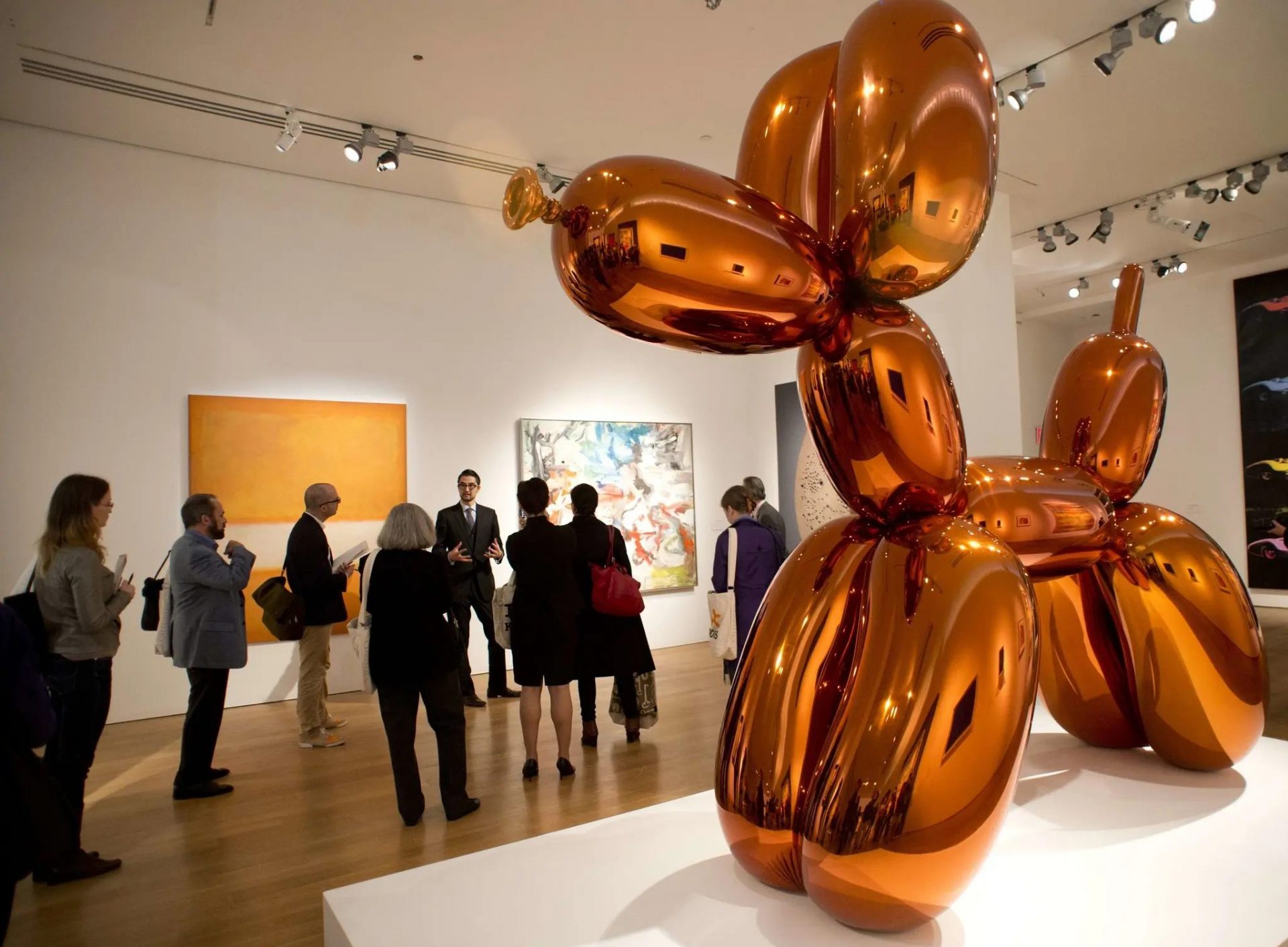 A photograph of an art gallery featuring a large orange sculpture in the shape of a ballon animal dog.