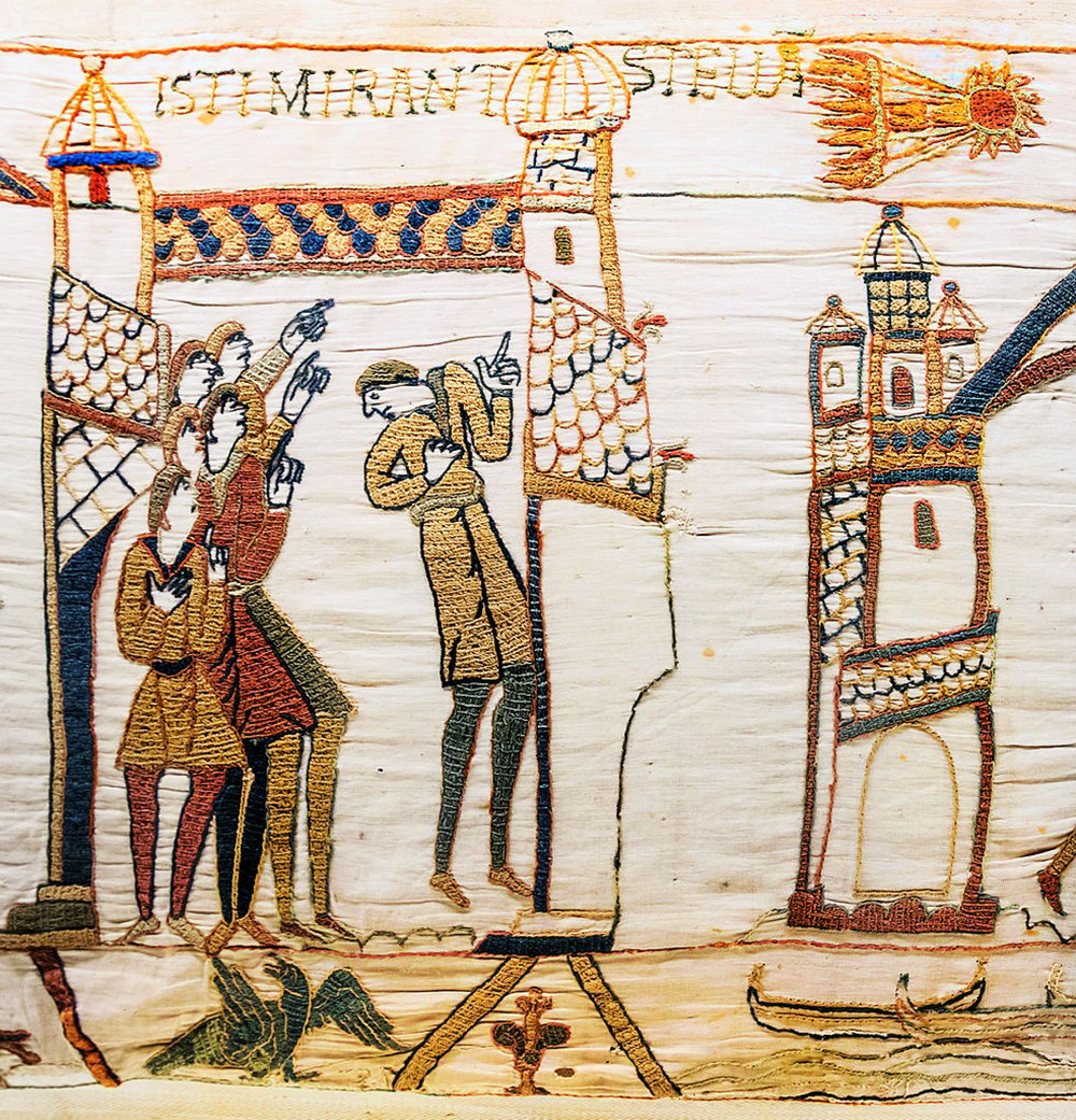 An image of the medieval Bayeux Tapestry tapestry made of beige fabric with colorful thread embroidery. The tapestry depicts men standing on a platform with a castle in the background. The men are looking at the Halley comet in the sky flying over a castle . The castle is red and white with blue flags on top.