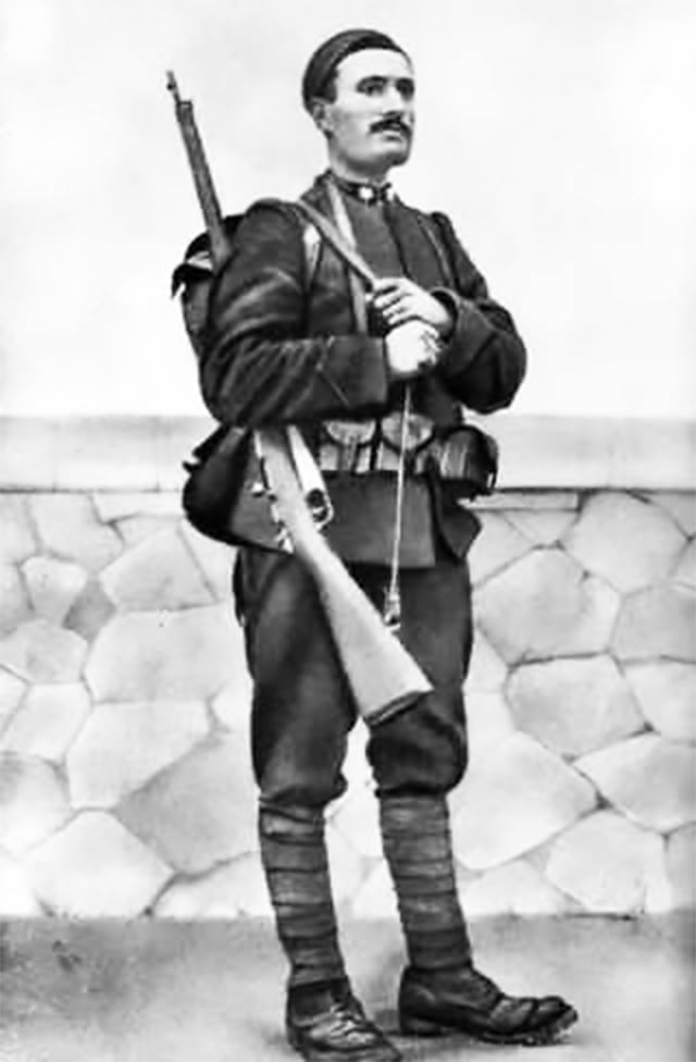 A historical photo of Benito Mussolini as a soldier posing with a rifle in front of a stone wall. The soldier is wearing a military uniform and a hat.