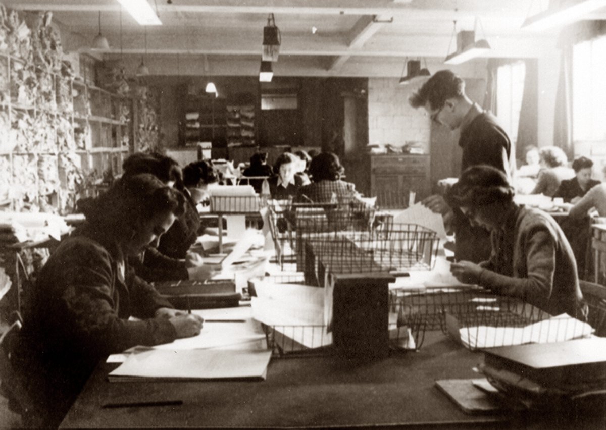 A black and white photo of a group of people working in a large room with high ceilings and large windows. The people are sitting at long wooden tables with papers and wire baskets on them. The people are working with their heads down, focused on their tasks.