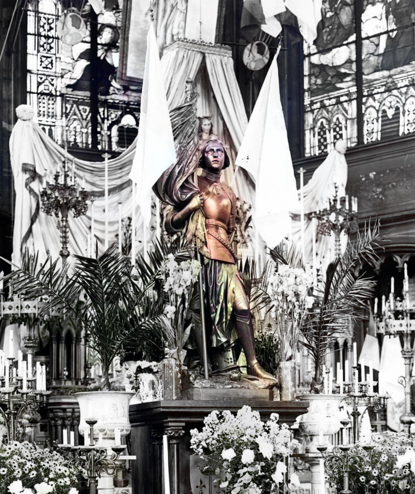 A black and white photo of a statue of Joan of Arc holding a banner at a church. The statue is on a pedestal surrounded by white flowers and candles. The background consists of a stained glass window and a large organ.
