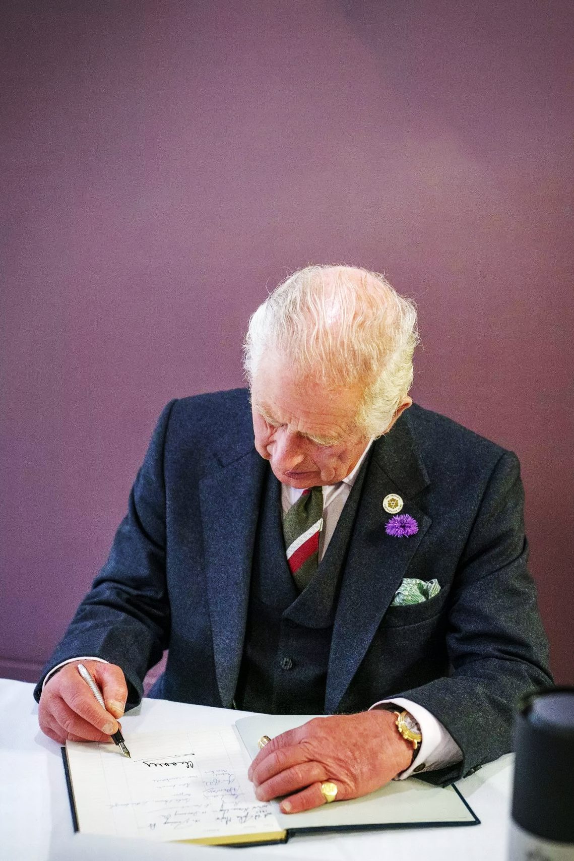 A photograph of King Charles sitting at a desk and signing a book using a fountain pen.