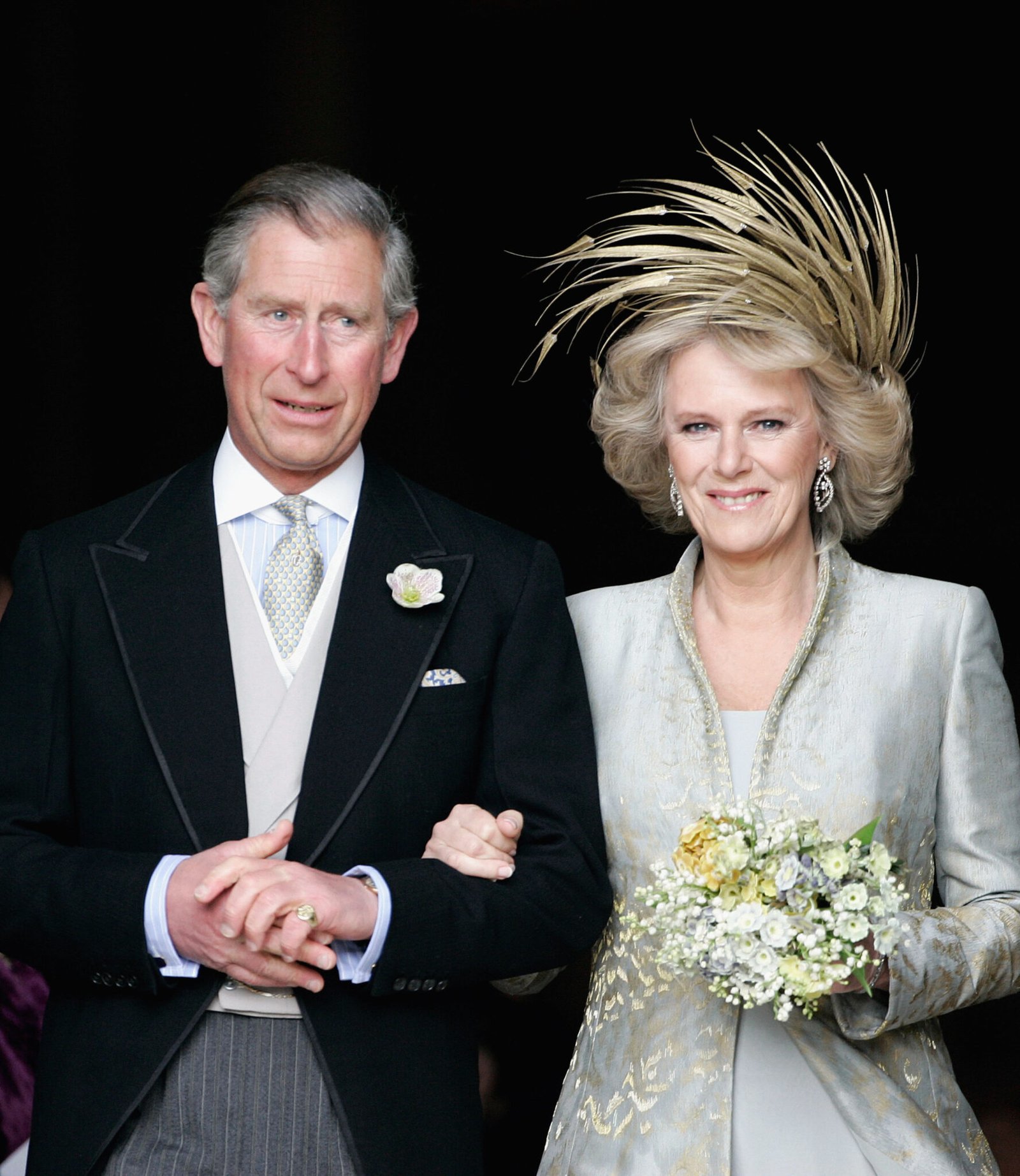 A photograph of the wedding of King Charles and Camilla Bowles, wearing elegant attire. Camilla has an elaborate hair piece made of feathers on and is holding a bouquet. Charles is wearing a tailcoat.