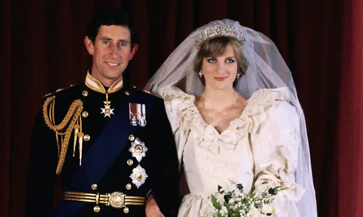 This is a photograph of Charles and Diana, the Prince and Princess of Wales, wearing wedding clothing and holding hands as they stand in front of a dark background. The prince is wearing a dress uniform, while the princess is wearing a white wedding dress and a tiara.
