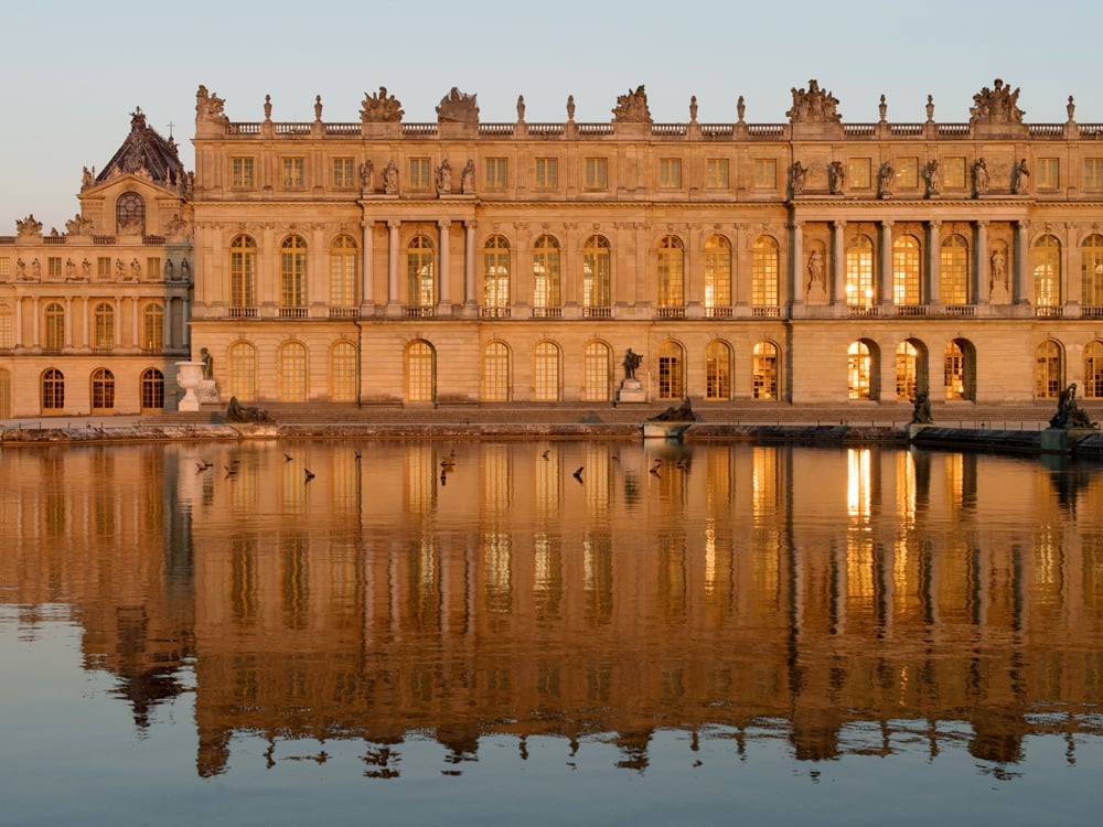 A photograph of Chateau of Versailles facade in the sunset, reflecting in water.