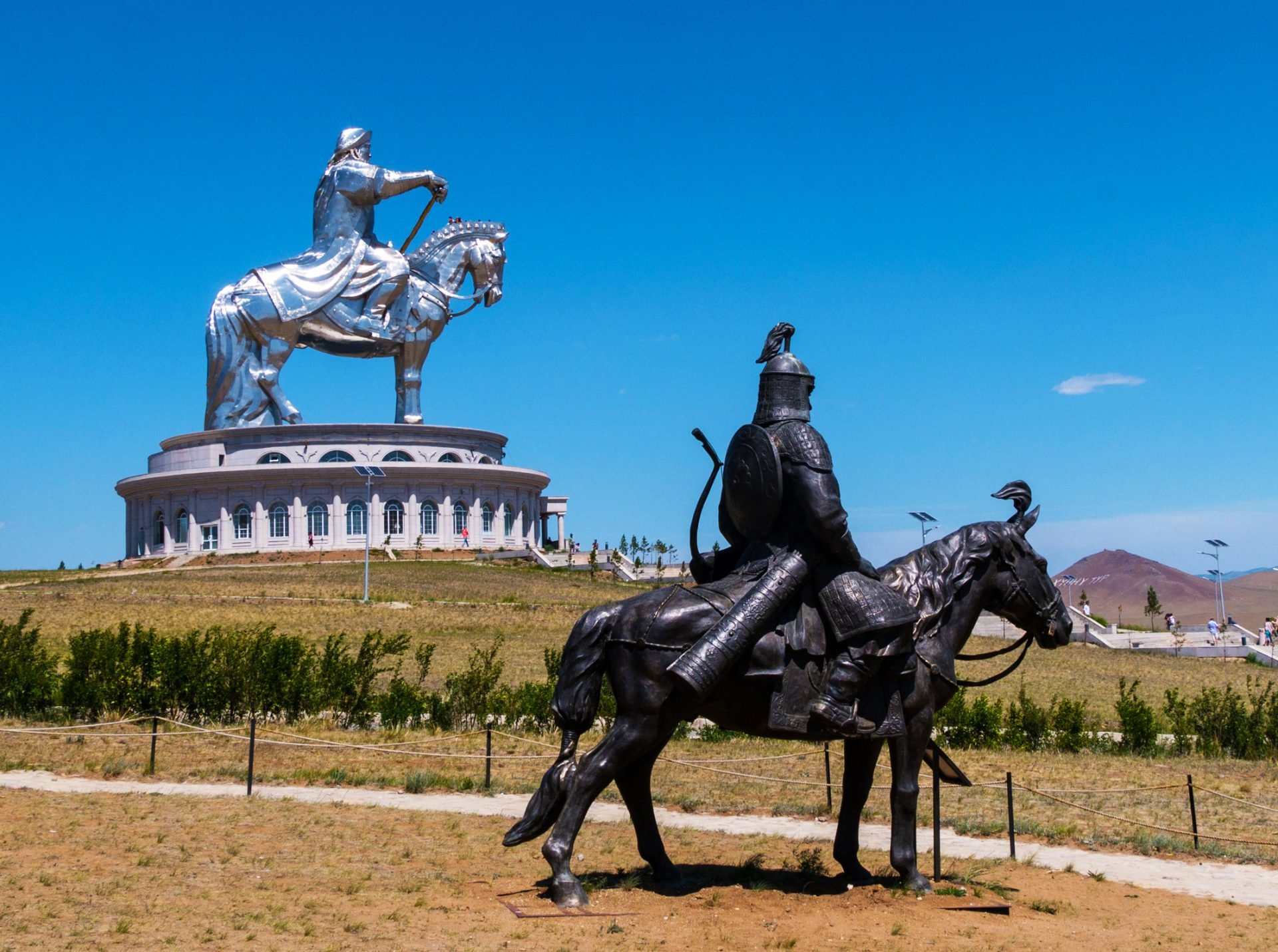 A photo of two statues of men on horses in a park in Mongolia. One statue is in the foreground and shows a man in armor with a sword and a rearing horse. The other statue is in the background and shows Genghis Khan, the world's largest 40-meter equestrian statue.