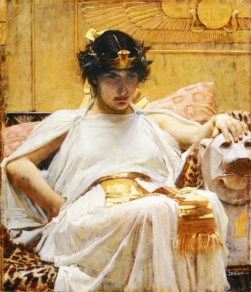 A painting of Cleopatra. She is depicted as sitting in a throne. She is wearing a light, white gown and a gold crown on her head. Her facial expression signals dissatisfaction.