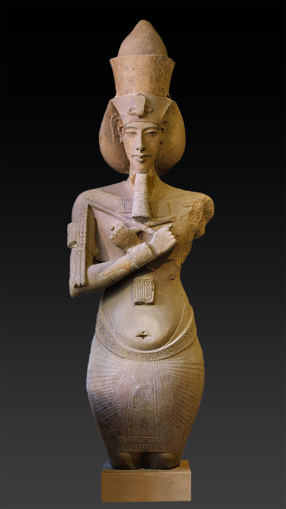 A stone statue of a standing figure of Amenhotep IV/Akhenaten. The statue is wearing a headdress and a skirt, and is on a square base. The background is black.