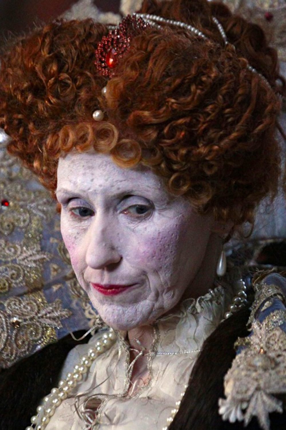 A photograph of a film still from Armada featuring a closeup of Queen Elizabeth. Her face is damaged by smallpox. She is wearing white makeup in an effort to conceal the damage.