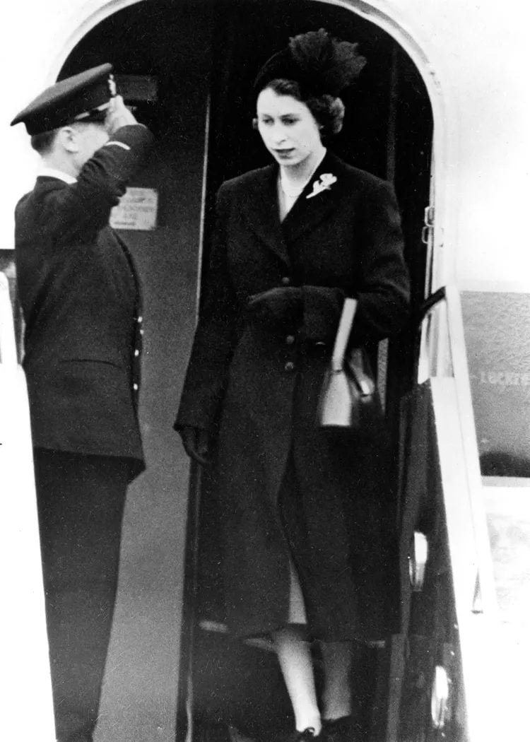 A black and white photograph of Queen Elizabeth II stepping out of a plane, dressed in black. An mad dressed in a uniform is standing next to her, saluting.