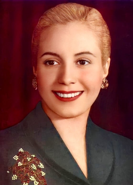 A colorized photograph of Evita Peron. She has blond hair slicked back, she looks happy and smiling. She is wearing a green top and elaborate jewelry.