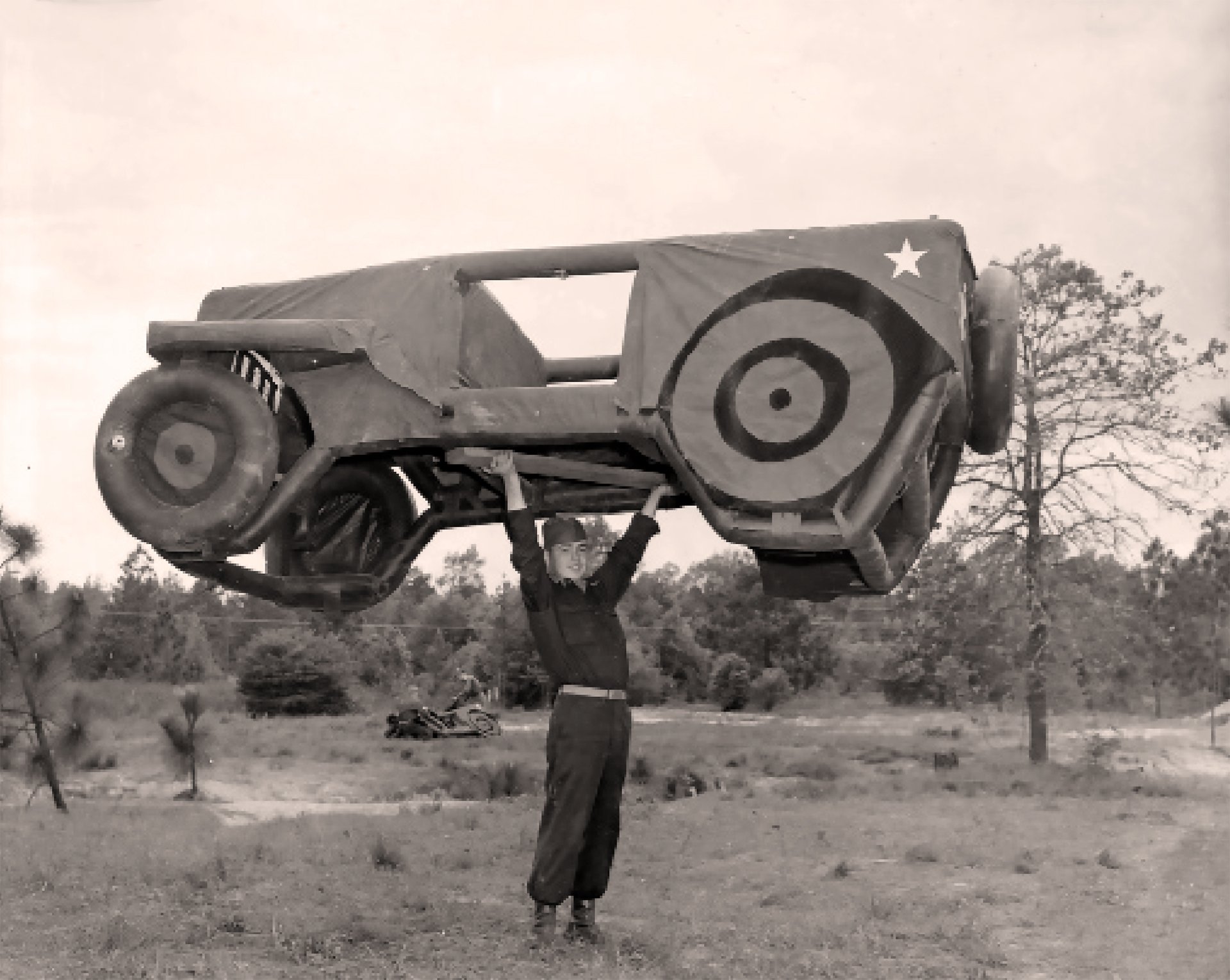 A black and white photo of a man lifting a large model of a military jeep over his head. The man is wearing a dark uniform and is standing in a field with trees in the background.