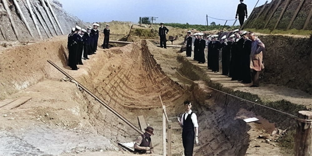 A colorized black and white photo of a group of people in suits and hats inspecting a large trench with a wooden support structure – excavation site of the ship burial at Sutton Hoo. The background is a barren landscape with a few trees and a hill.