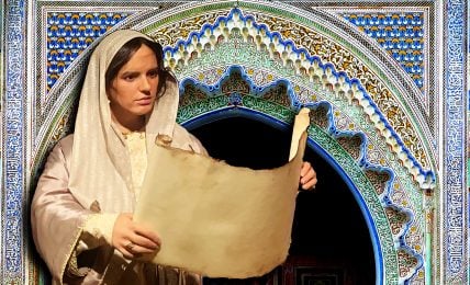 A museum exhibit of a Fatima Al-Fihri mannequin in a white robe holding a parchment scroll in front of a blue and white tiled archway.