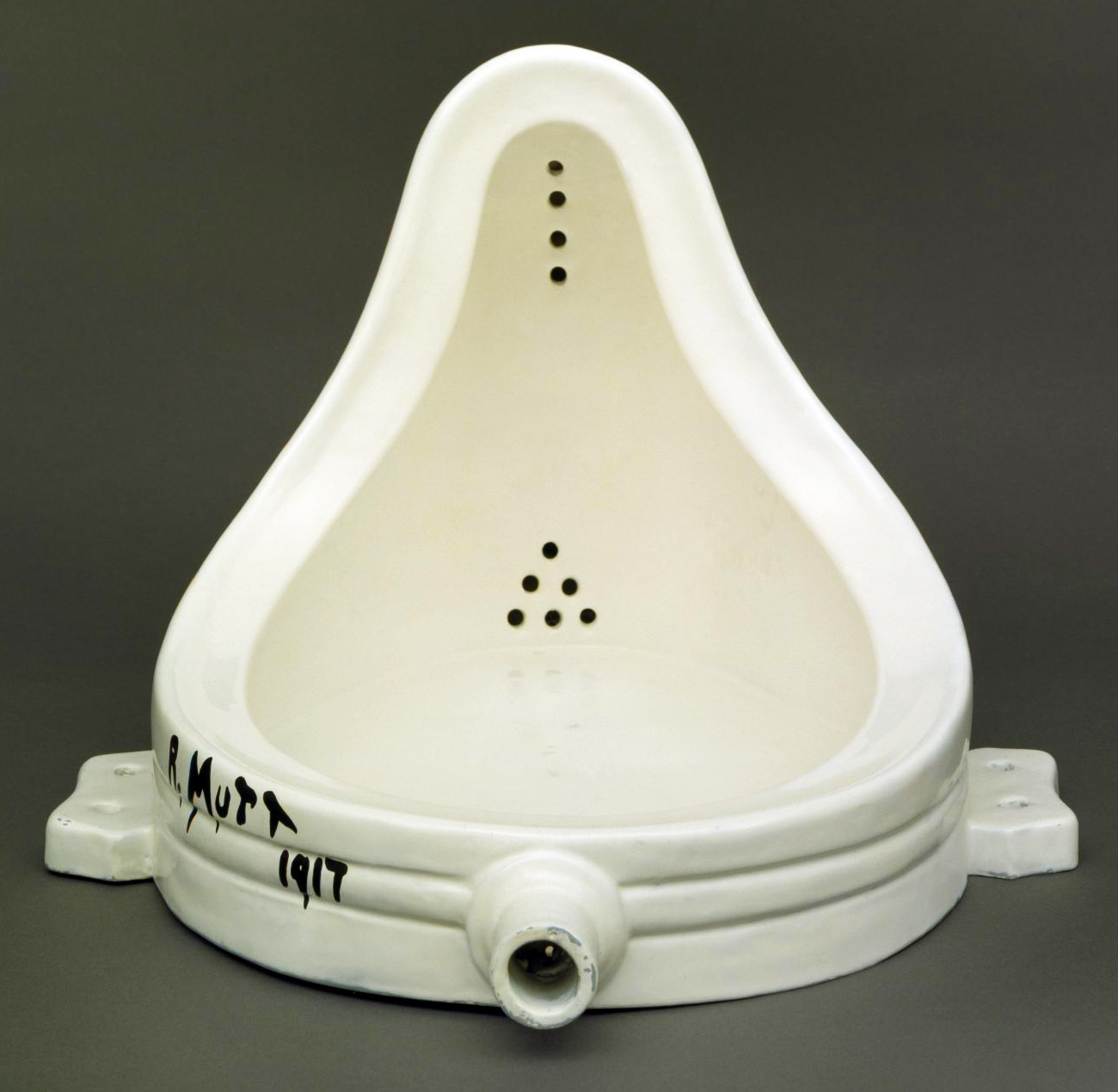 White porcelain urinal turned artwork, titled 'Fountain' by Marcel Duchamp. The urinal is signed 'R. Mutt 1917' in black paint on its side. The piece is positioned against a dark gray background and showcases its curvilinear form and drain holes.