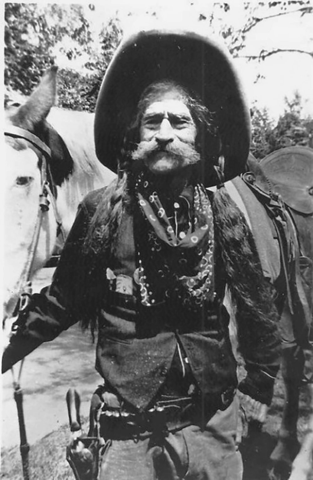 This is a black and white photograph of a cowboy in a western outfit. He has a large prominent mustache. He is wearing a cowboy hat, a dark shirt, and dark pants. He is standing next to a white horse. The horse is wearing a bridle and a saddle. In the background, trees are visible.