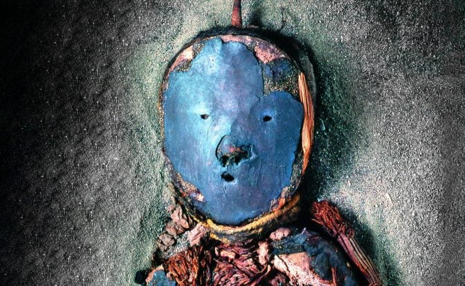 A close up photograph of the head of a mummy, wearing a blue mask.