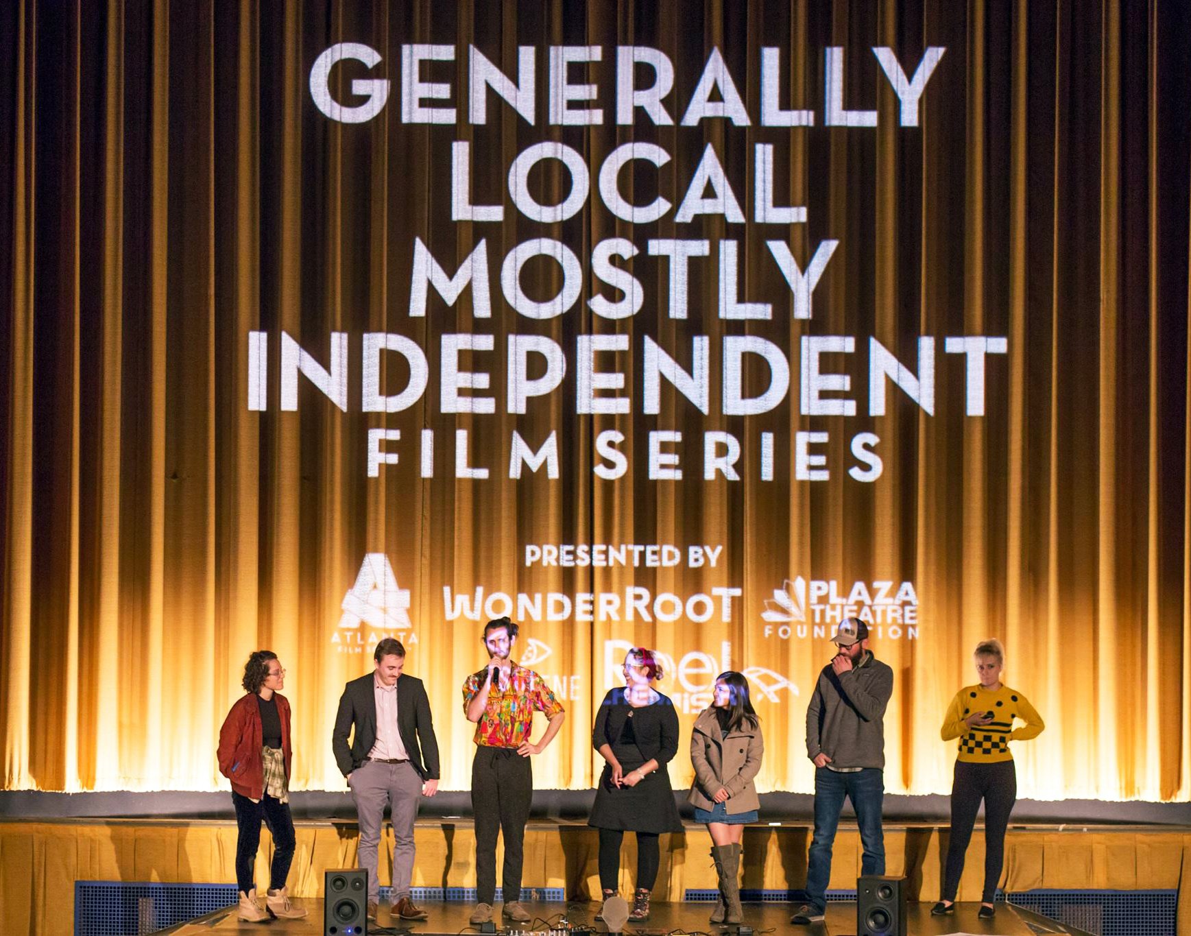 A group of people standing on a stage in front of a large screen that says “Generally Local Mostly Independent Film Series”. The people are facing the audience and wearing casual clothes. The stage is lit with yellow and orange lights.
