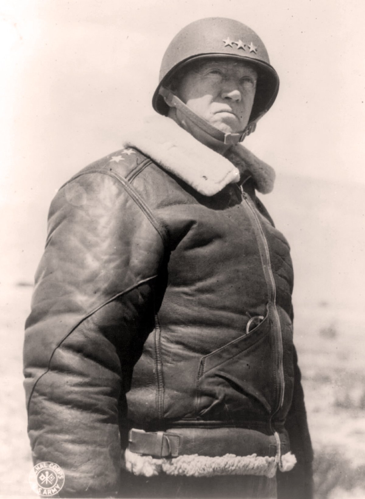 A black and white photo of General George Patton wearing a leather jacket and a helmet. The helmet is a dark color with three white stars. The jacket is a bomber style with a fur collar.