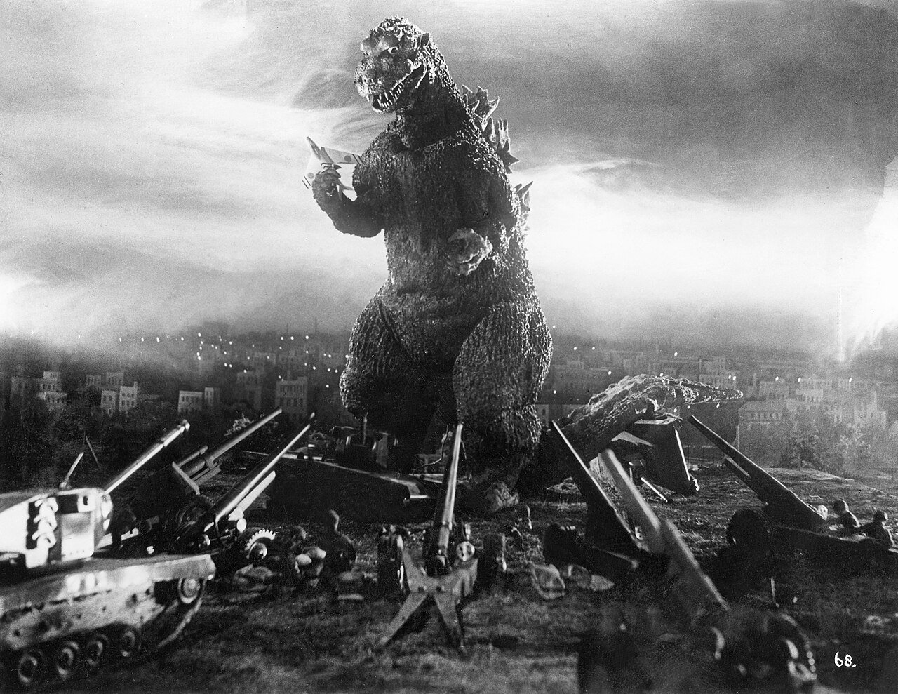 Godzilla holding a fighter jet in his hand and being fired upon by tanks