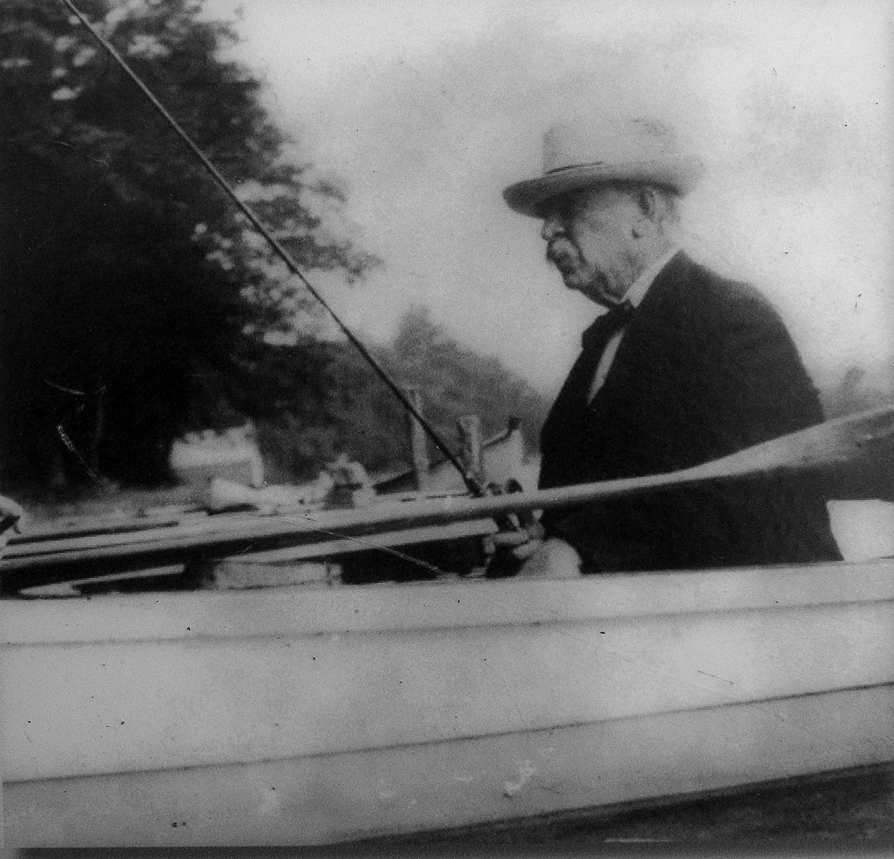 In this black and white image, a man in a suit and hat is seen sitting in a boat. He is holding a fishing rod in his hands. He looks determined as he stares ahead, his face partially visible in profile.