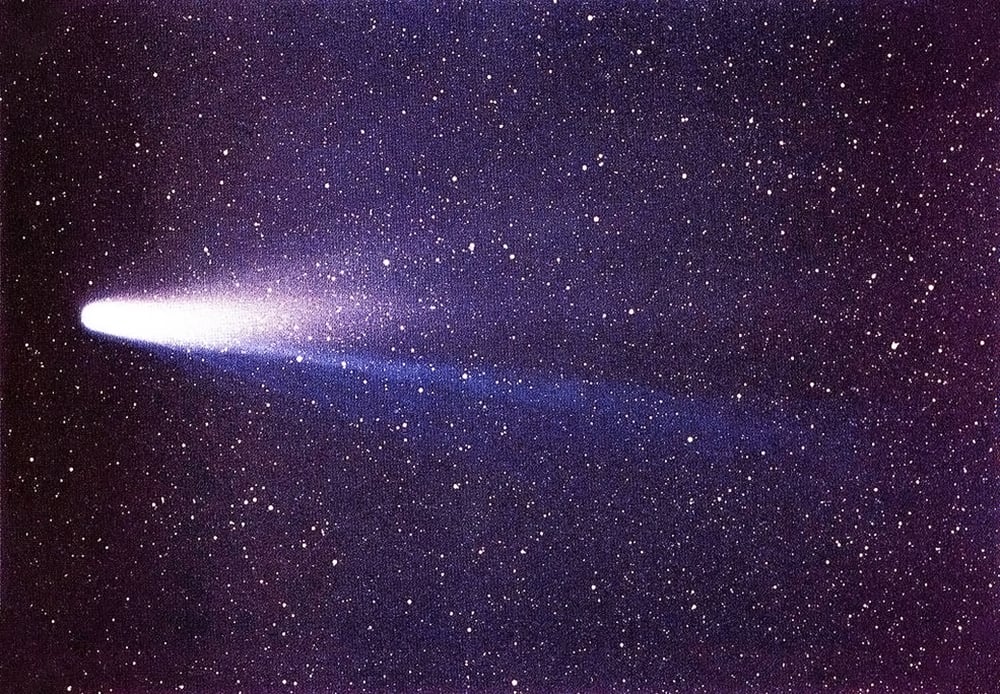 A photo of a Halley comet streaking across a starry night sky. The comet has a bright white head and a glowing tail. The background is a deep purple with many small white stars scattered throughout. The image is taken from a perspective that makes the comet appear large and close to the viewer.