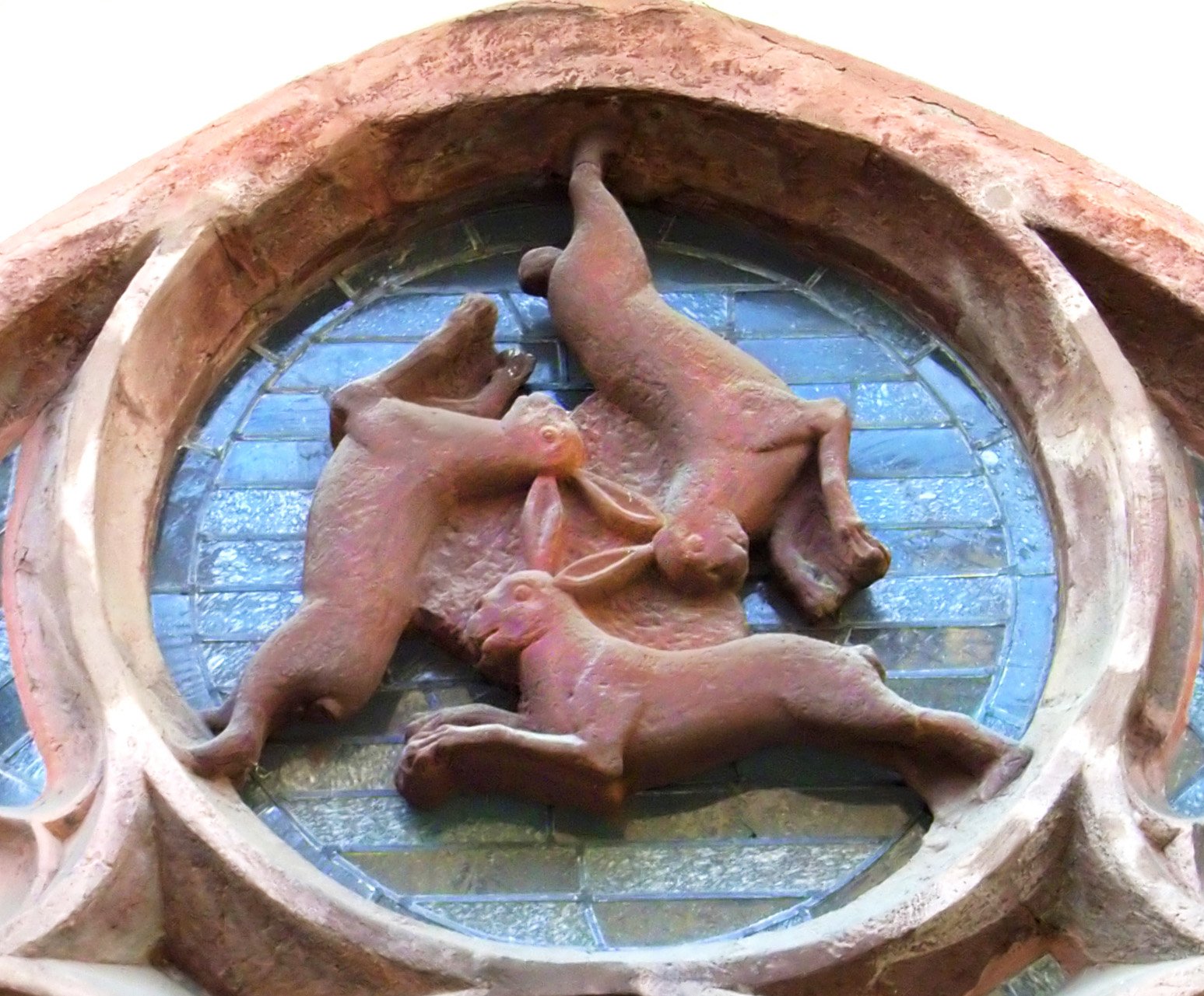 A terracotta sculpture of three rabbits in a circle, set in a round window with a blue background and a brick wall. The rabbits are playful and intertwined, their three ears joined together to form a triangle between them depicting the Holy Trinity.