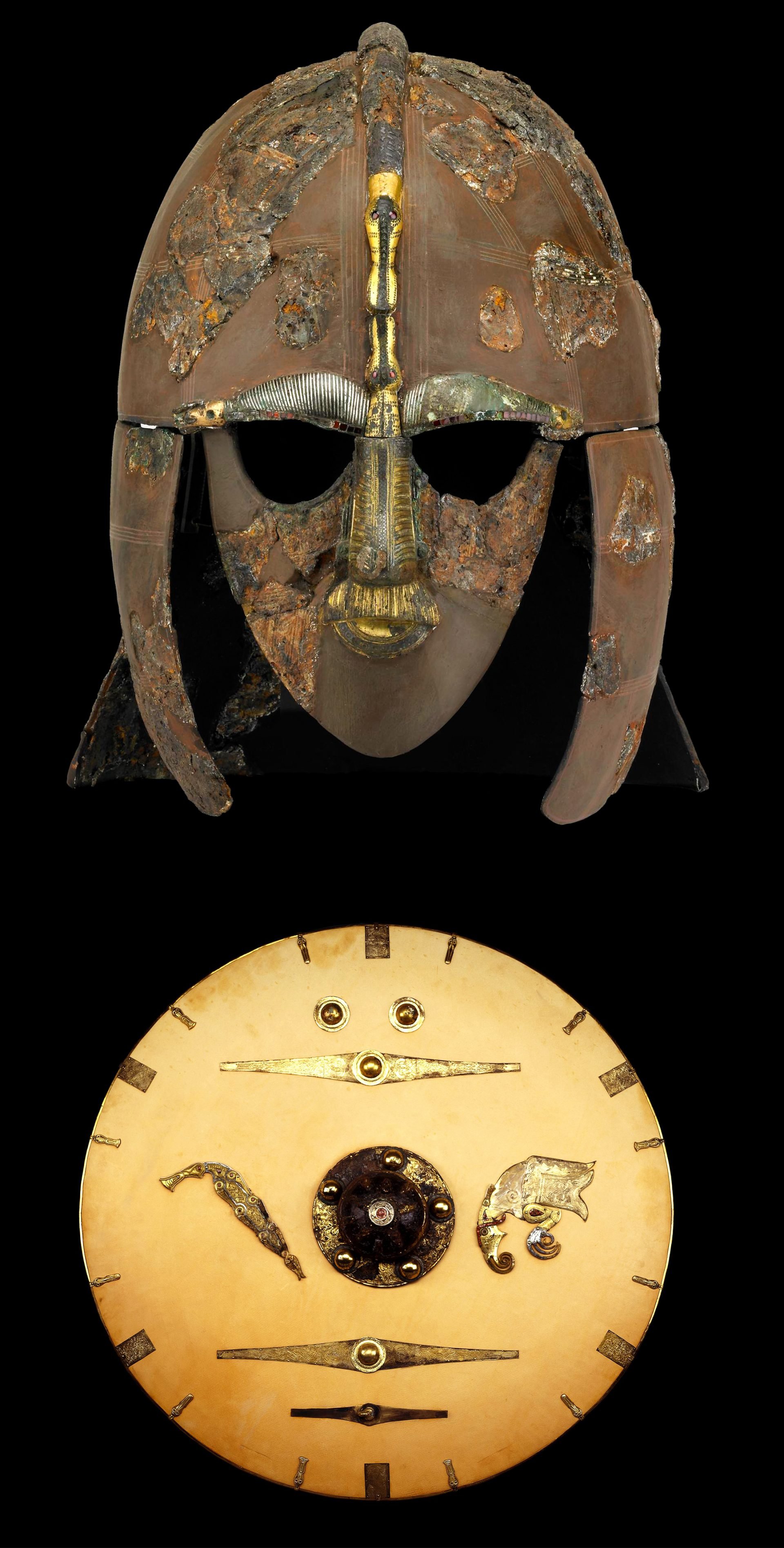 Restored iron helmet adorned with tinned copper alloy sheets that form a cap, cheek-pieces, mask, and neck-guard. The copper sheets showcase stamped designs of intertwined animals and warrior motifs. Adjacent is a Sutton Hoo shield replica, incorporating original elements of gold, garnet, copper alloy, and iron, alongside modern fittings on a lime wood base.