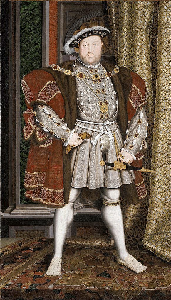 This portrait depicts king Henry VIII, a man wearing a 16th century-style garment. He is standing in an indoor setting, with the walls and floor of the room painted brown. The man has dark hair and appears to be in his late thirties or early forties. He is wearing a long-sleeved shirt that buttons up at the front, along with white tights and shoes of matching color. On top of this he wears a waistcoat with intricate detailing on it, including embroidery around the edges and pockets as well as decorative buttons down its length. His outfit is completed by a hat which sits atop his head, adding to the overall Tudor era look of his ensemble.