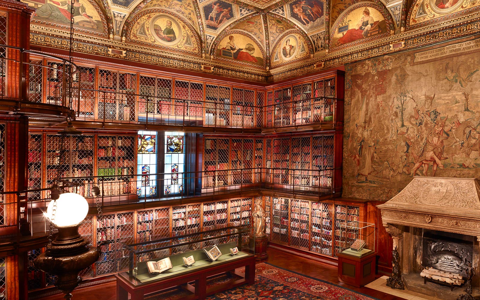 This is a beautiful library, with a high ceiling and ornately decorated walls and ceiling. The bookshelves are lined with red and gold painted wood, and are filled with books of all sizes. The room is filled with light from the large centrally placed window. There is a large fireplace in the center of the room.