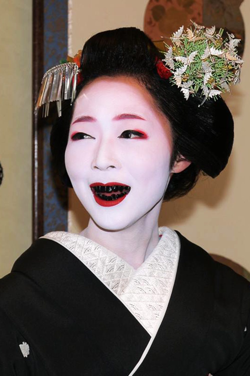 The woman in the image is wearing a black and white kimono with white lace and a white flower on her head. She has long black hair and black eyes. She has black teeth and white facial makeup.
