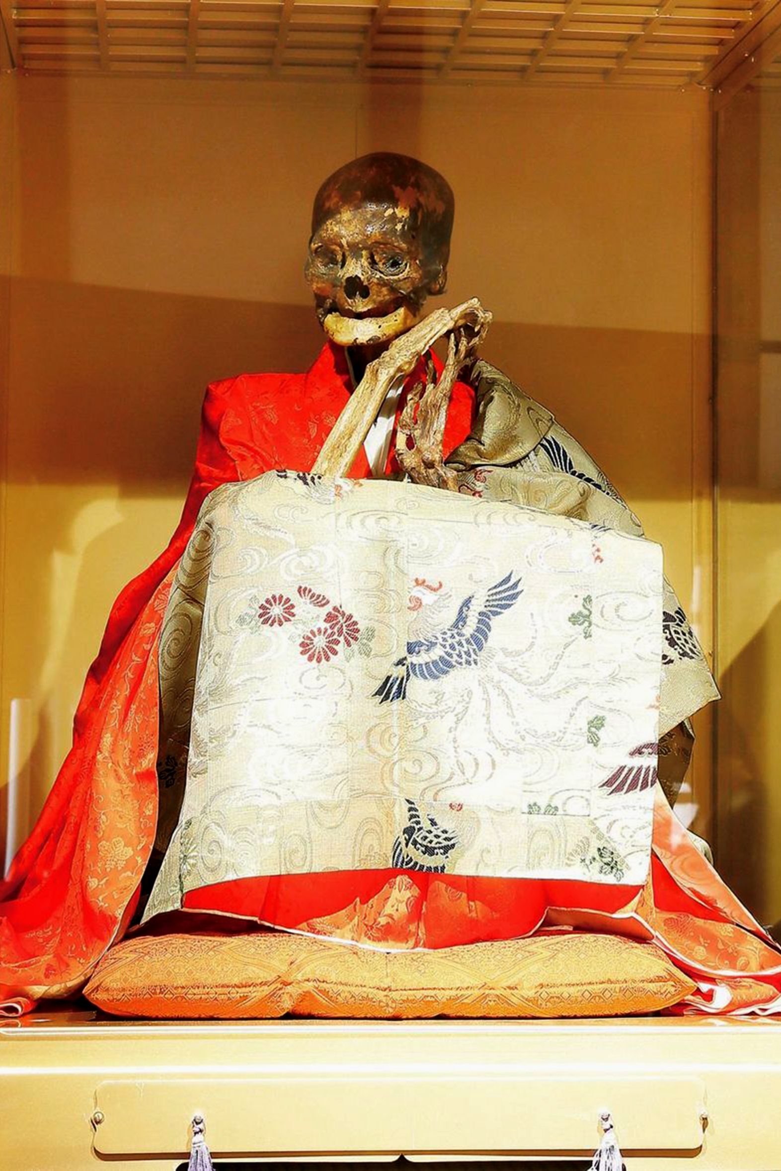 A photograph of a mummified Japanese monk. The mummy is in a sitting position inside a glass cabinet. It is wearing a bright red robe.