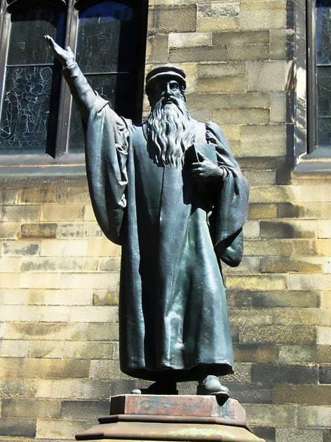 Bronze statue of a bearded man - John Knox - in robes, holding a book in one hand and pointing upwards with the other. The statue stands in front of a stone building of New College with stained glass windows.