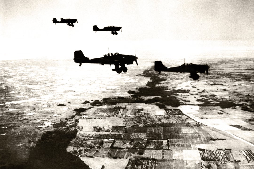 A black and white photo of a group of four Junkers Ju-87 “Stuka” dive-bombers flying over a landscape of fields and farmland.