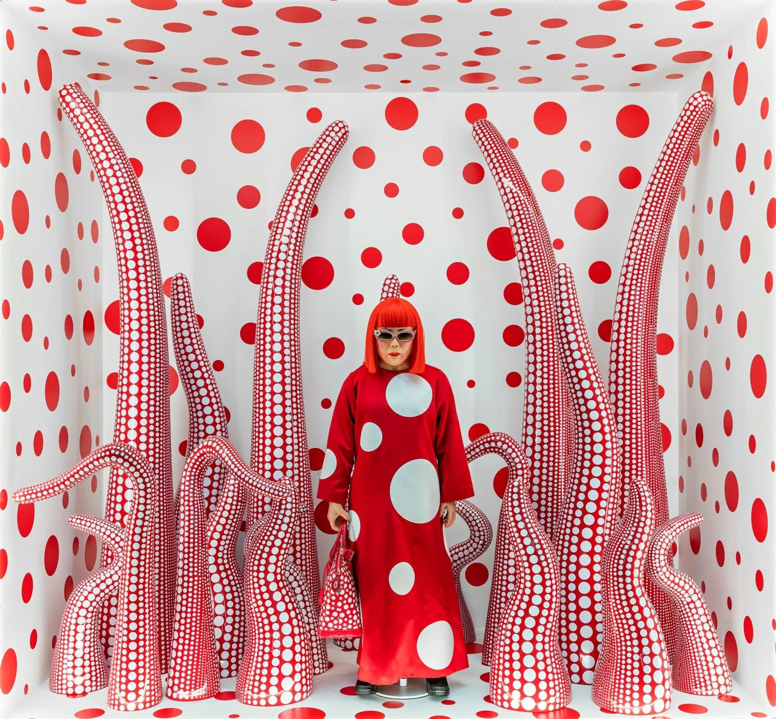 A woman in a red wig and a red dress with large white circles is standing inside a box. She is surrounded by large tentacles protruding from the floor.
