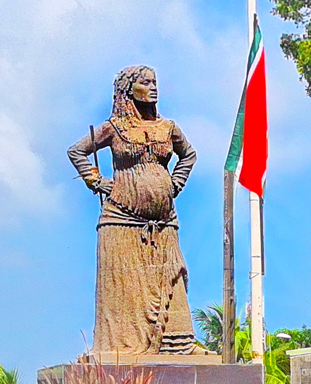 A statue of Mulatresse Solitude on a pedestal next to a flagpole with a flag of Guadeloupe. She is pregnant and is wearing a dress.