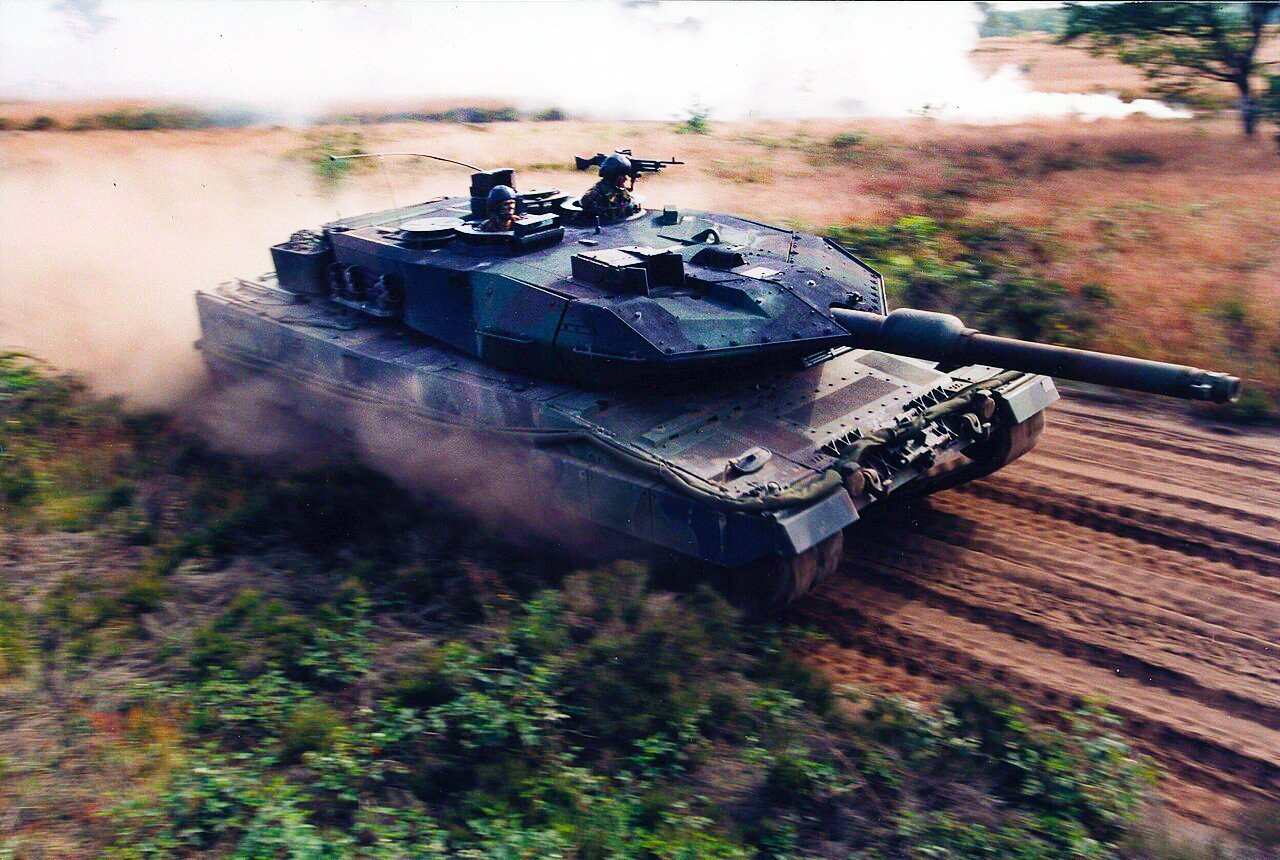 A photo of a Leopard 2A5 battle tank moving quickly through a field with dust trailing behind it.