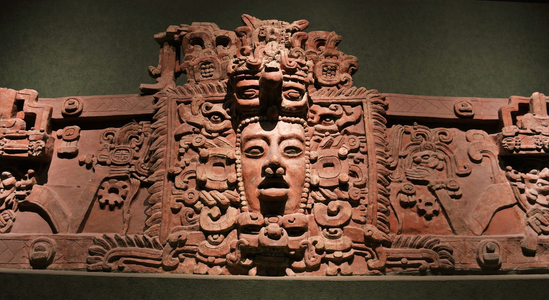 A photo of an ancient Maya temple decoration stone sculpture of a face with intricate details and patterns. The sculpture is in a museum setting with a green background.