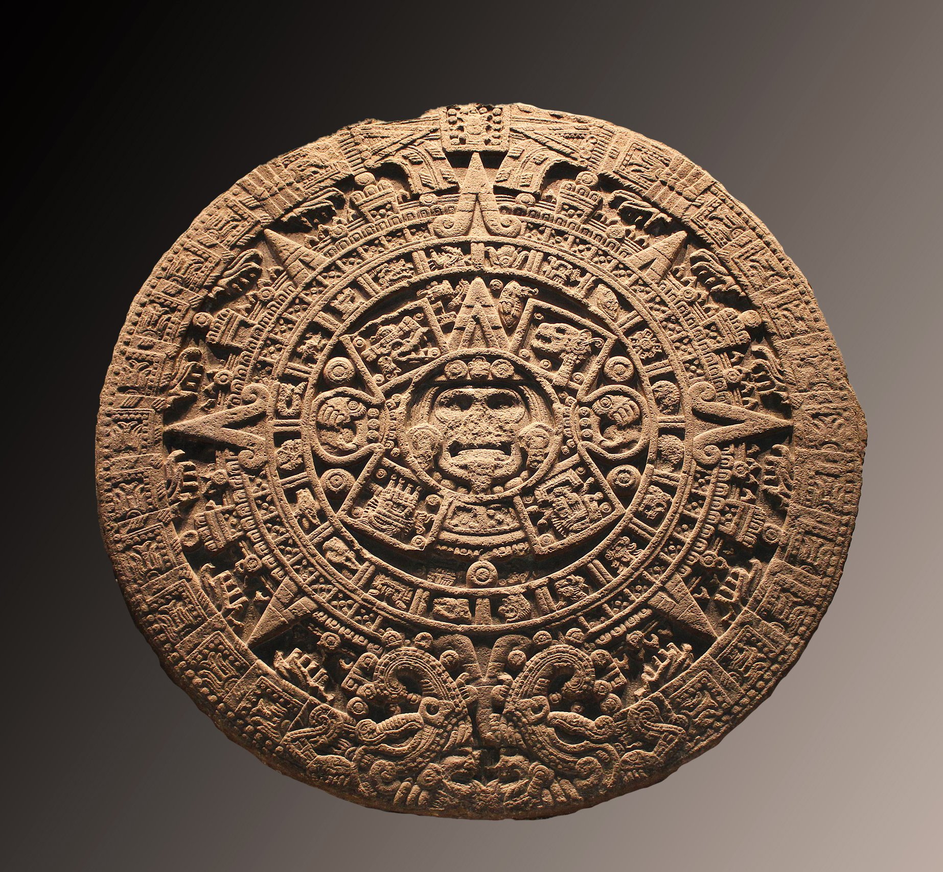 A photo of an ancient Mayan calendar stone. The stone is circular in shape and has intricate carvings on it. The center of the stone has a face with a tongue sticking out. The outer ring of the stone has various symbols and figures carved into it. The stone is a brownish color and appears to be made of a type of rock or clay. The background is black, which makes the stone stand out.