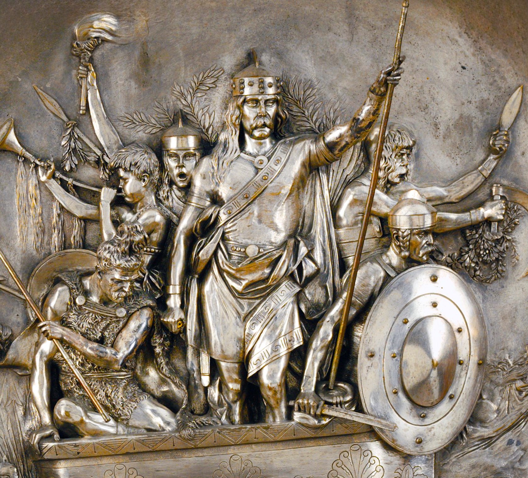 A metallic sculpture of a group of warriors in silver and gold tones, set against a background of foliage. The central warrior king Merovech holds a spear, while other smaller warriors hold spears, shields and swords.