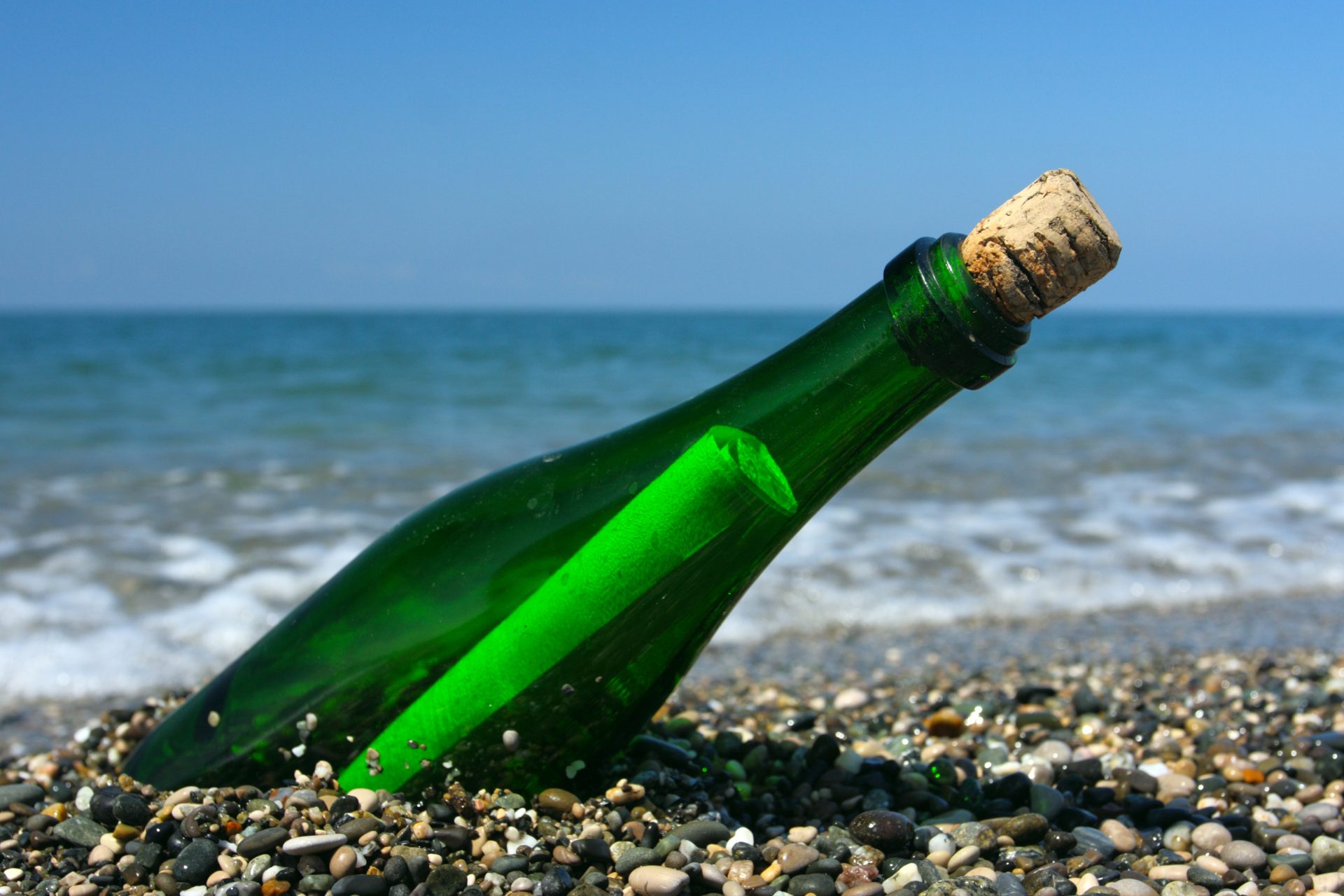 A photograph of a rolled up piece of paper in side a green bottle, half buried in pebbles on a rocky beach.