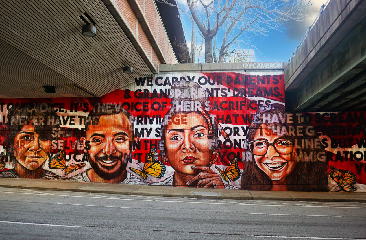 A photo of a mural on a brick wall with four faces of people. The mural is in red, white and black colors with orange butterflies. The mural reads “We carry our grandparents’ dreams and the voice of their sacrifices. We may not have their battle lines but we have their imagination.” The mural is signed by the artist “@ycambron”.
