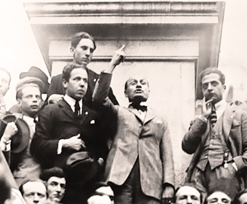 Benito Mussolini pointing his finger in the sky in the act of speaking, he is surrounded by people in suits and ties during the first Fascist rally in Rome, Italy, in 1919