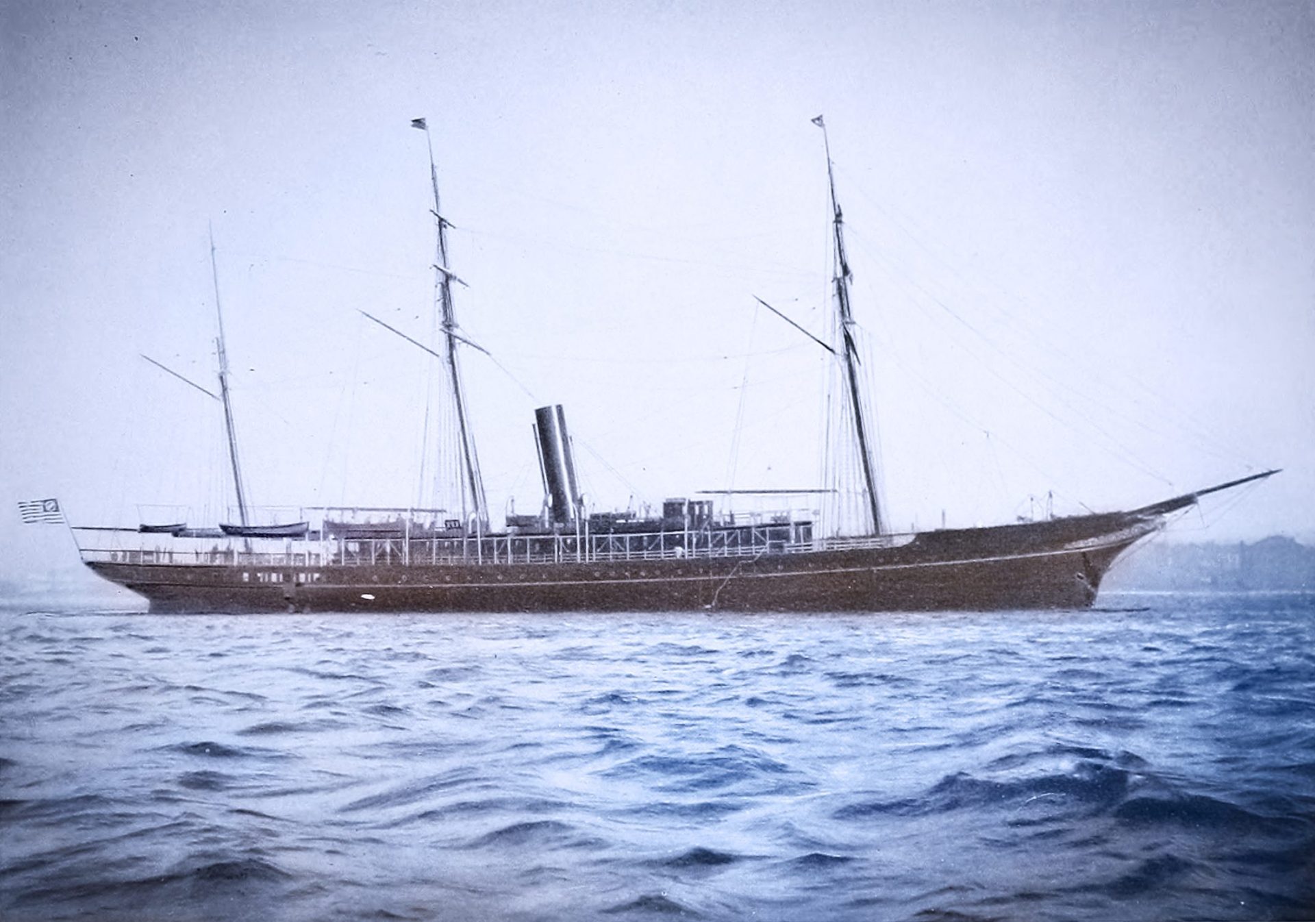The photo shows a 19th century yacht in the middle of a large body of water. The yacht is very large and has three tall masts. The hull is dark and appears to be made of wood.