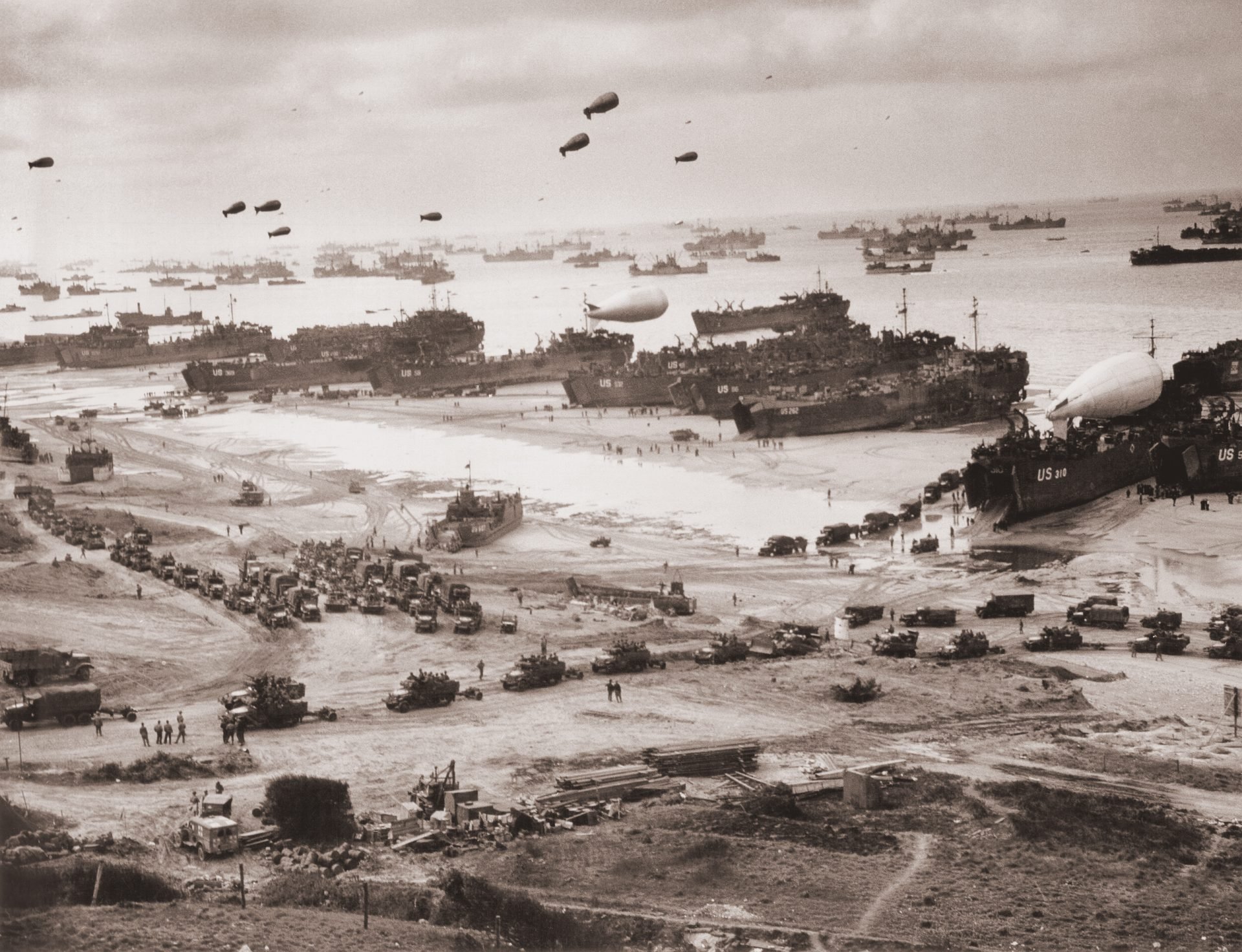 A historical photograph of a beach landing during a military operation, showing the beach and the sea filled with military vehicles, equipment, personnel, ships and boats. There are also blimps and parachutes in the sky. The photograph is black and white, grainy and faded.