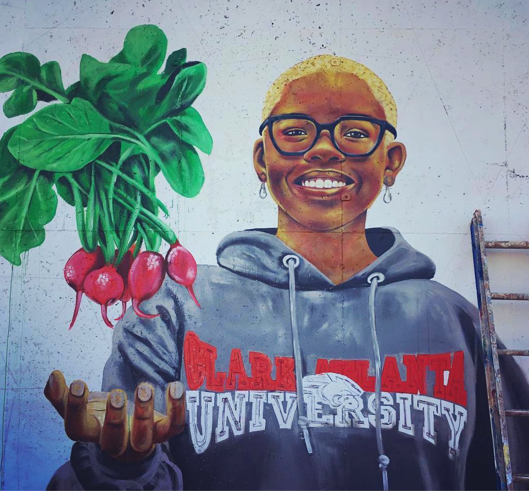 A mural of a woman in glasses in a gray hoodie with Clark Atlanta University logo. A bunch of radishes are landing into her hand.